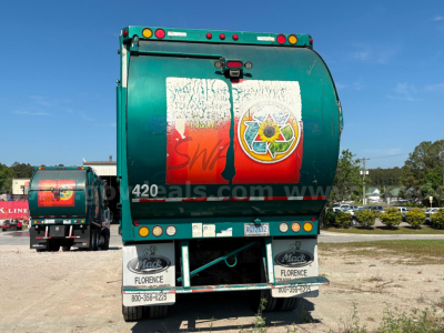 Rear view of a green Mack MRU613 refuse truck showing curbside bumper and waste container with logo and taillights.
