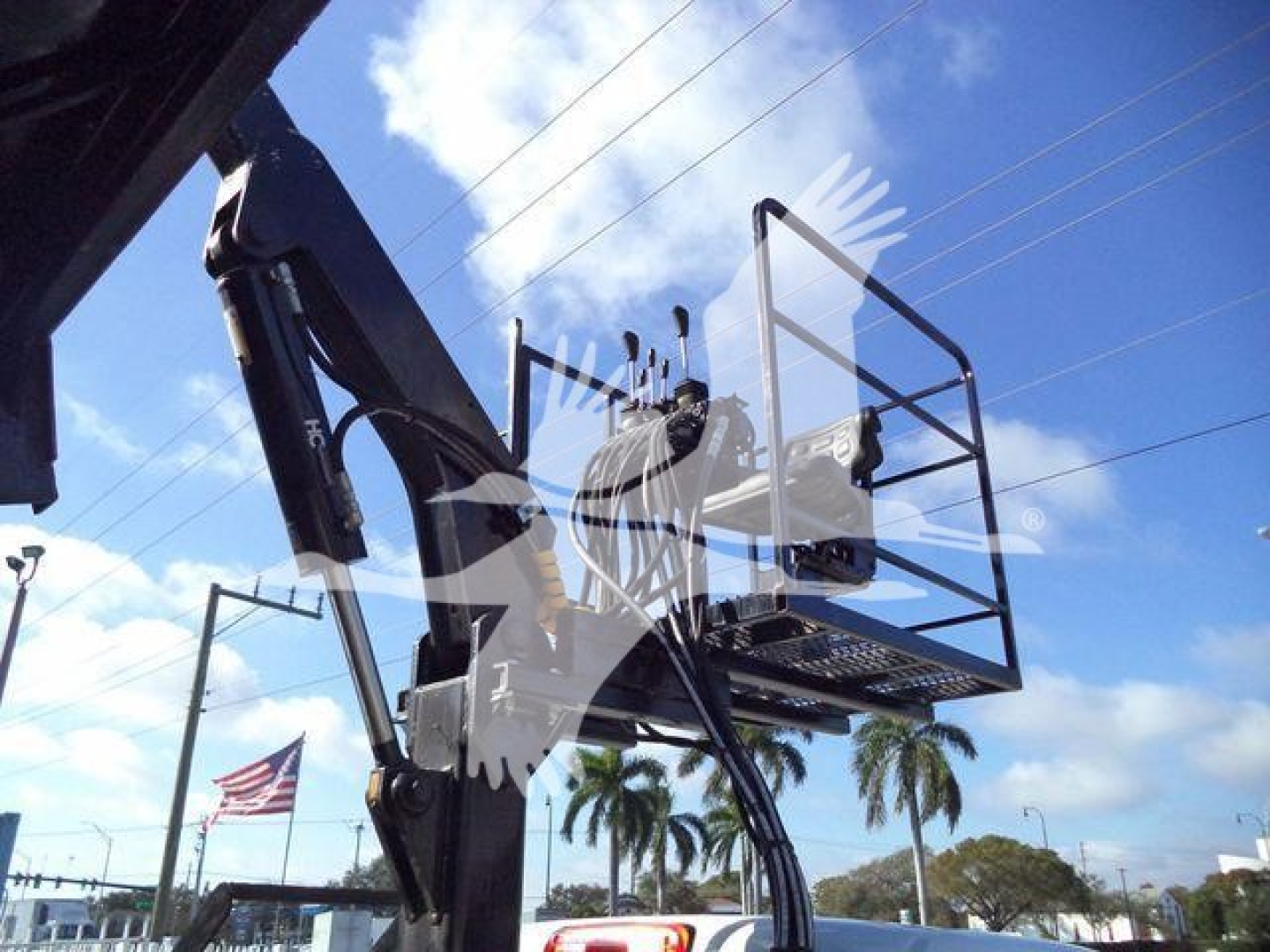 Truck chassis or bucket lift with platform in view, hydraulic arms and metal basket visible against blue sky with palm trees.