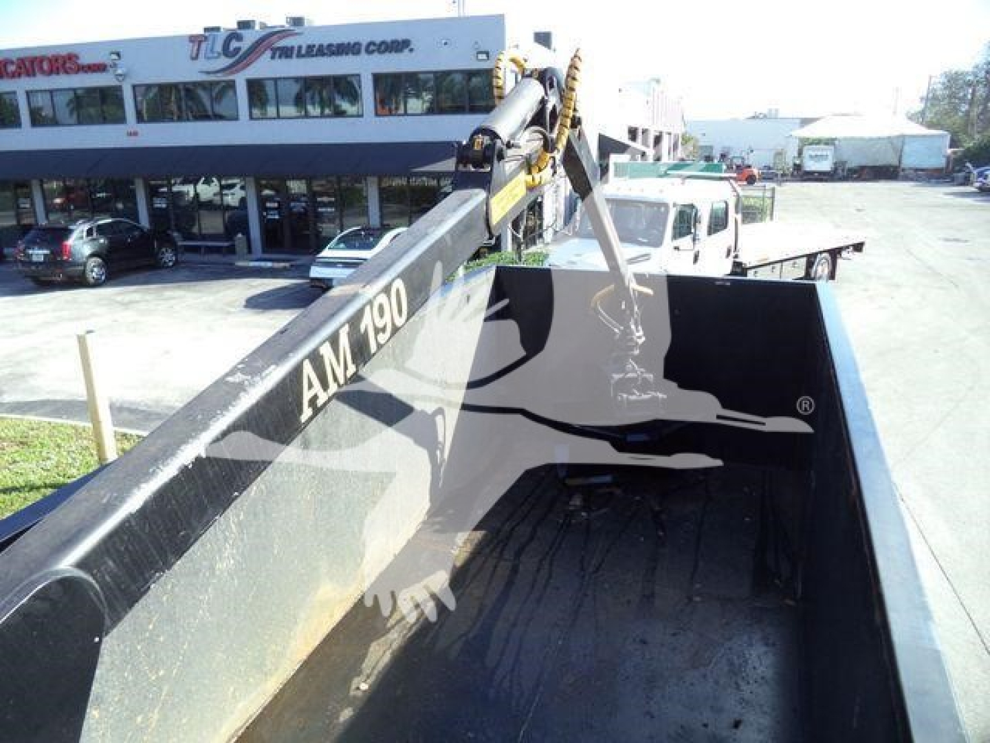Truck bed view of a Chevrolet Silverado 6500HD with a hydraulic dump chute, black bed interior, and a crane arm labeled AM 790. Background shows dealership building.