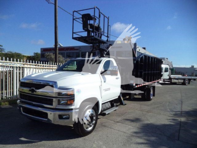 2023 Chevrolet Silverado 6500HD dump truck in a dealership lot with a lift platform on the rear; front grille with Chevrolet emblem, white cab, dual rear wheels, and equipment bed.