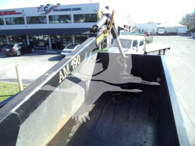 Truck bed view of a Chevrolet Silverado 6500HD with a hydraulic dump chute, black bed interior, and a crane arm labeled AM 790. Background shows dealership building.