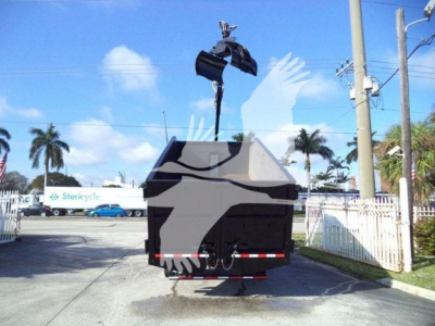 Truck bed with a hydraulic grapple attachment lifting a large container; street with palm trees in background. Visible text includes Stericycle on a distant vehicle.