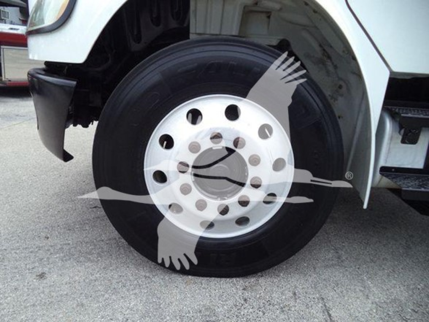 Front wheel of a Freightliner heavy truck showing a polished steel wheel with multiple circular holes and a black tire.