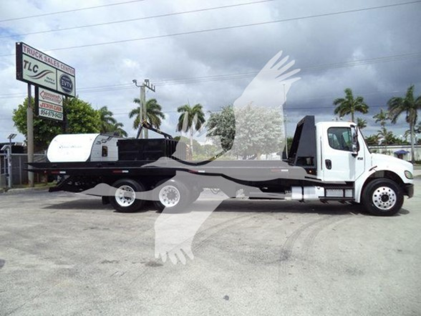 Flatbed tow truck with a white cab and long lowbed trailer, Freightliner Business Class M2 rollout, palm trees in background.