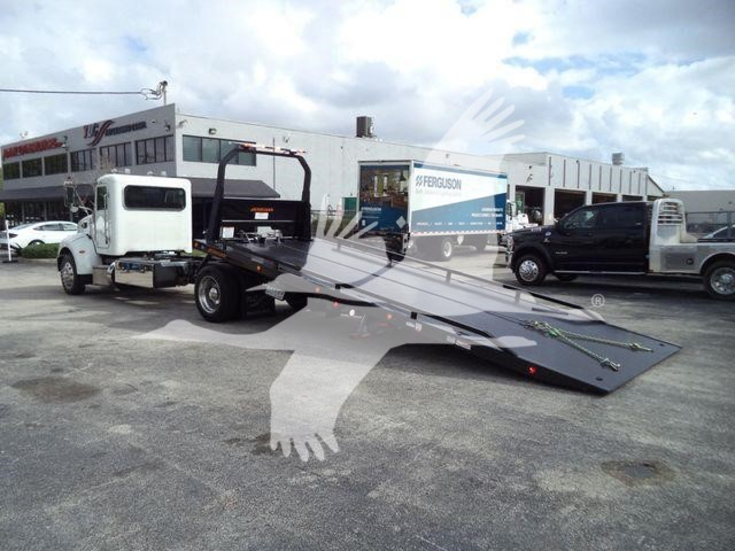Peterbilt 337 rollback tow truck with flatbed extended, white cab, in a dealership lot with other vehicles in the background.