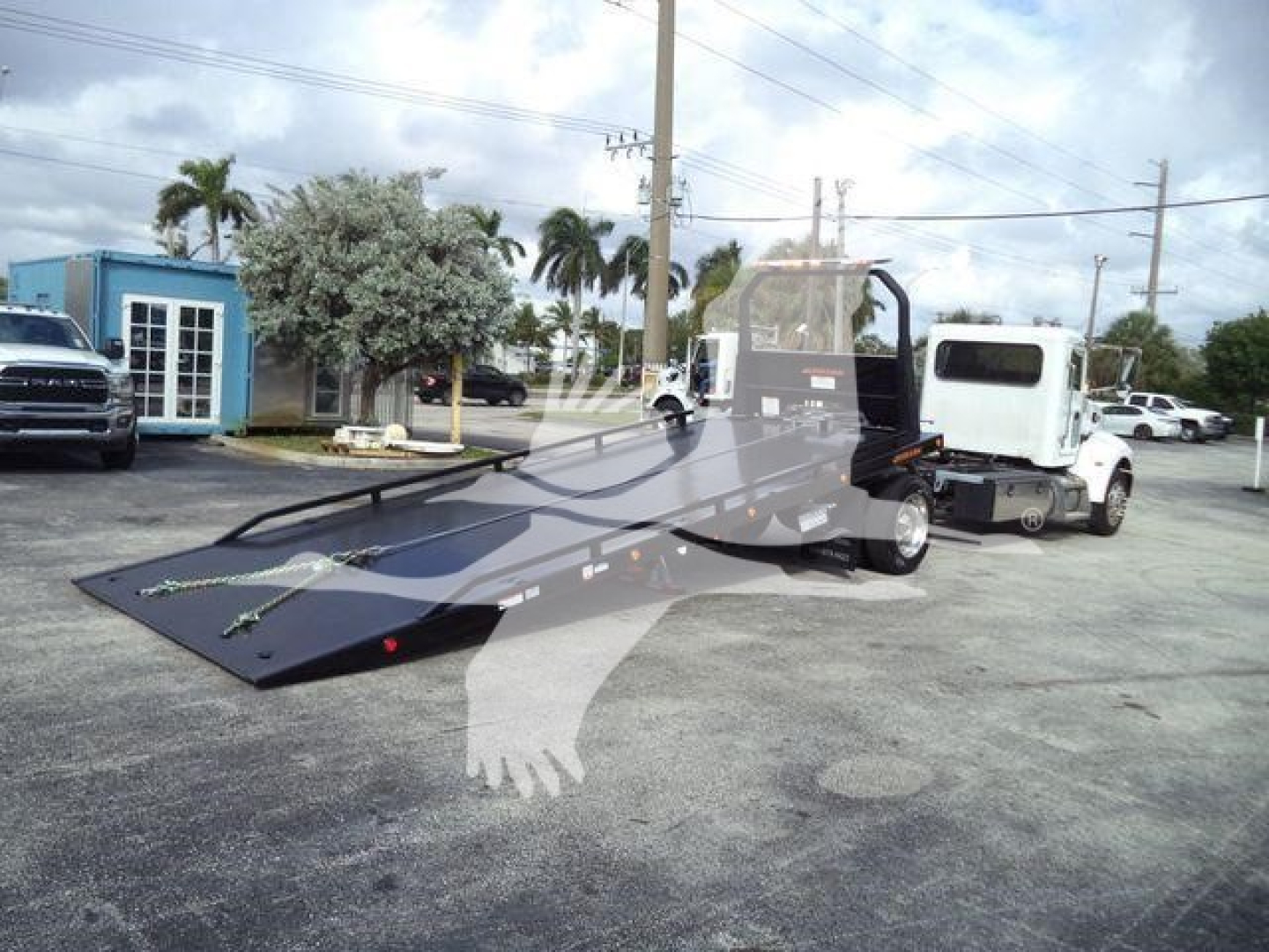 Peterbilt 337 rollback tow truck with flatbed in service showing front cab and hydraulic bed angled, with another white truck in background.