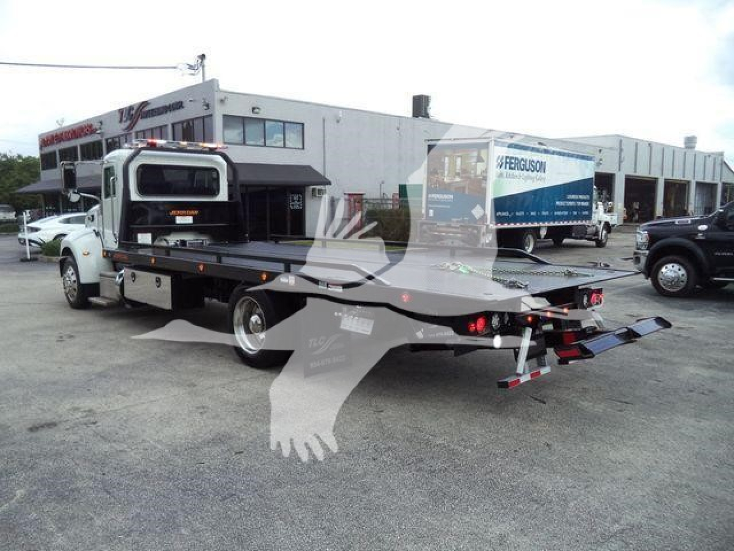 Flatbed tow truck in a lot with a white cab and a black flatbed; in background a building with trucks and signs; no legible model text visible on the truck.
