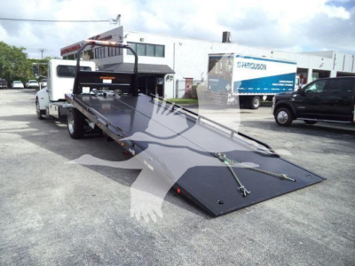 2019 Peterbilt 337 rollback tow truck with a lowered flatbed extended toward the ground; visible text on the truck includes no readable labels, background shows another truck with 'Ferguson' branding.