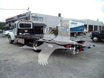 Flatbed tow truck in a lot with a white cab and a black flatbed; in background a building with trucks and signs; no legible model text visible on the truck.
