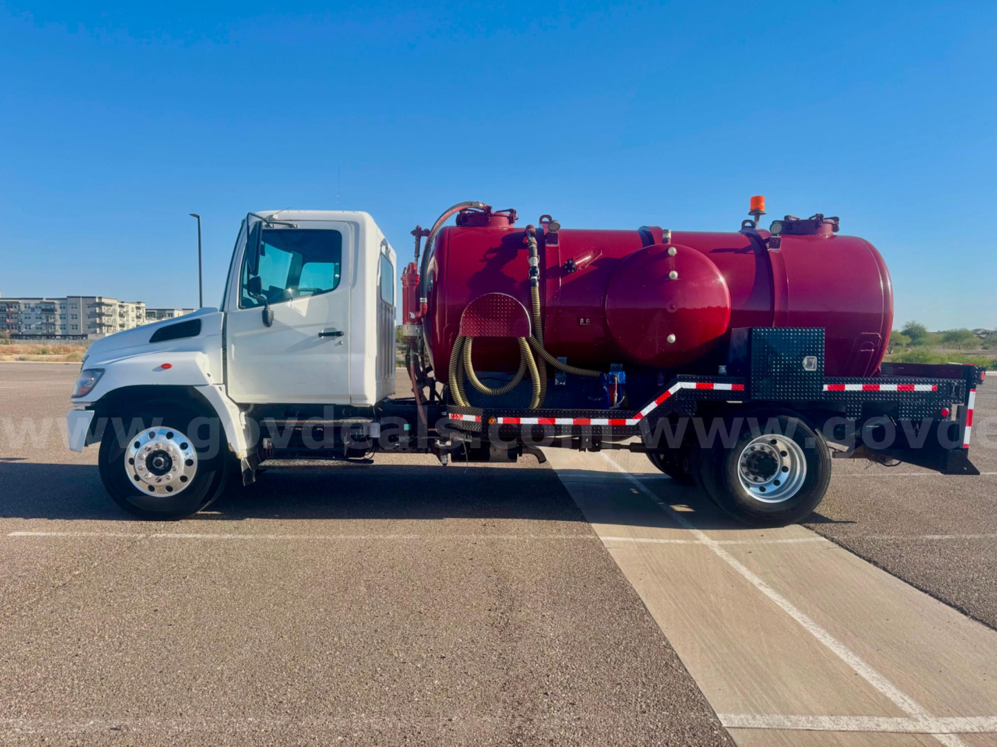 Truck-mounted vacuum tank unit on a white cab chassis, red cylindrical tank with hoses and controls, side view