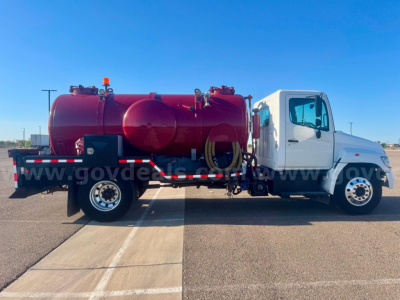 Hino vacuum tank truck side view showing large red cylindrical tank on flatbed with white cab and various hoses and controls.