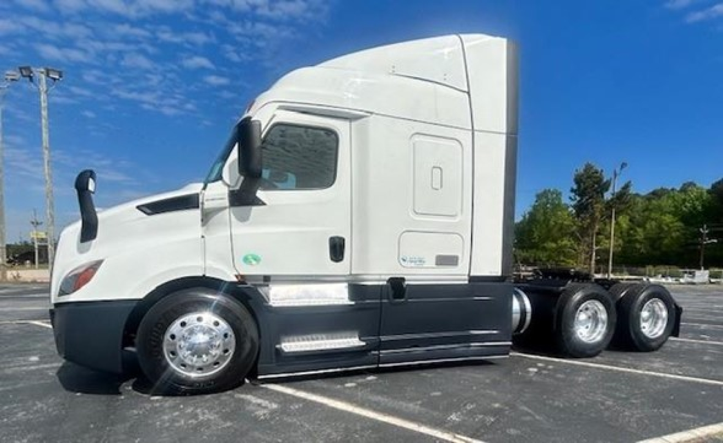 Freightliner Cascadia 116 sleeper tractor in a parking lot, side profile view, with white cab and black lower body, visible wheels and chrome fuel tanks.
