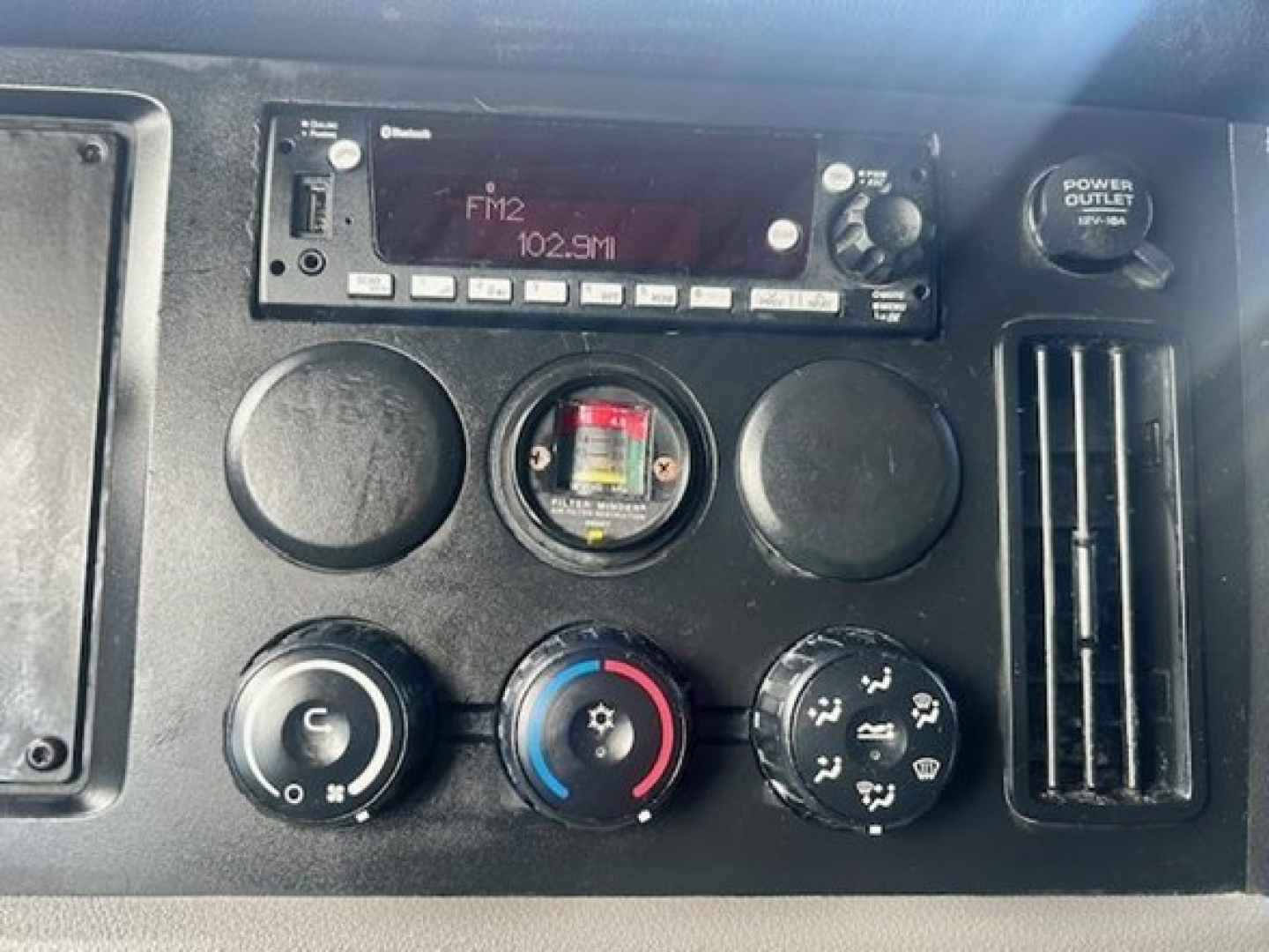Dashboard of a Freightliner Cascadia shows FM2 audio display ‘102.9M’ and climate controls including circular vents, dials, and a power outlet.