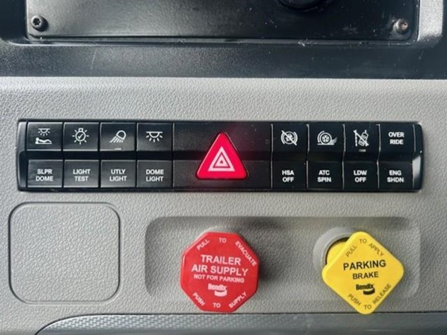 Freightliner Cascadia dashboard switches showing hazard button, trailer air supply red knob, and parking brake yellow knob with labels.