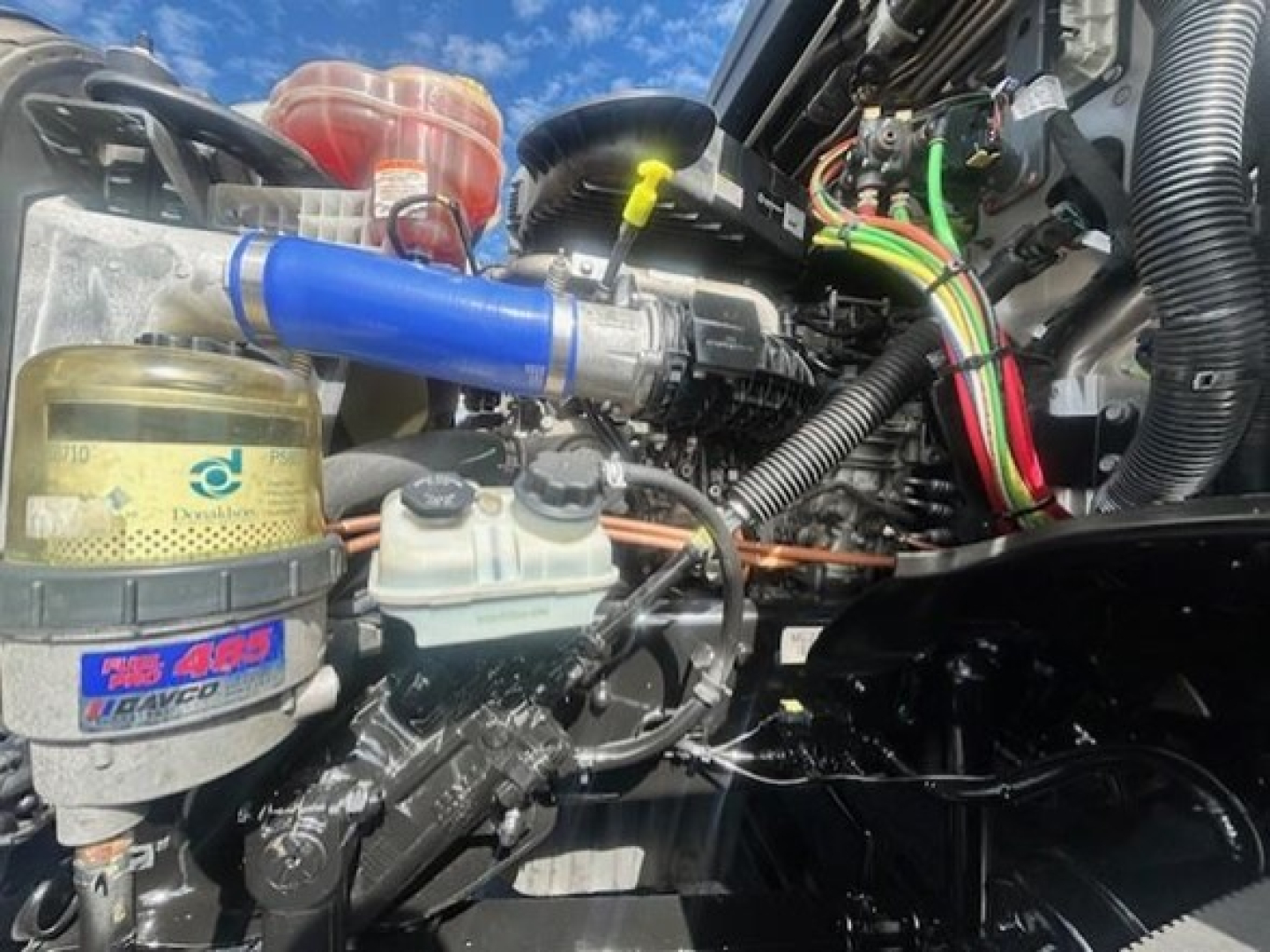 Freightliner Cascadia engine bay with blue silicone hose, oil and coolant reservoirs, wiring harnesses, and accumulator nearby.