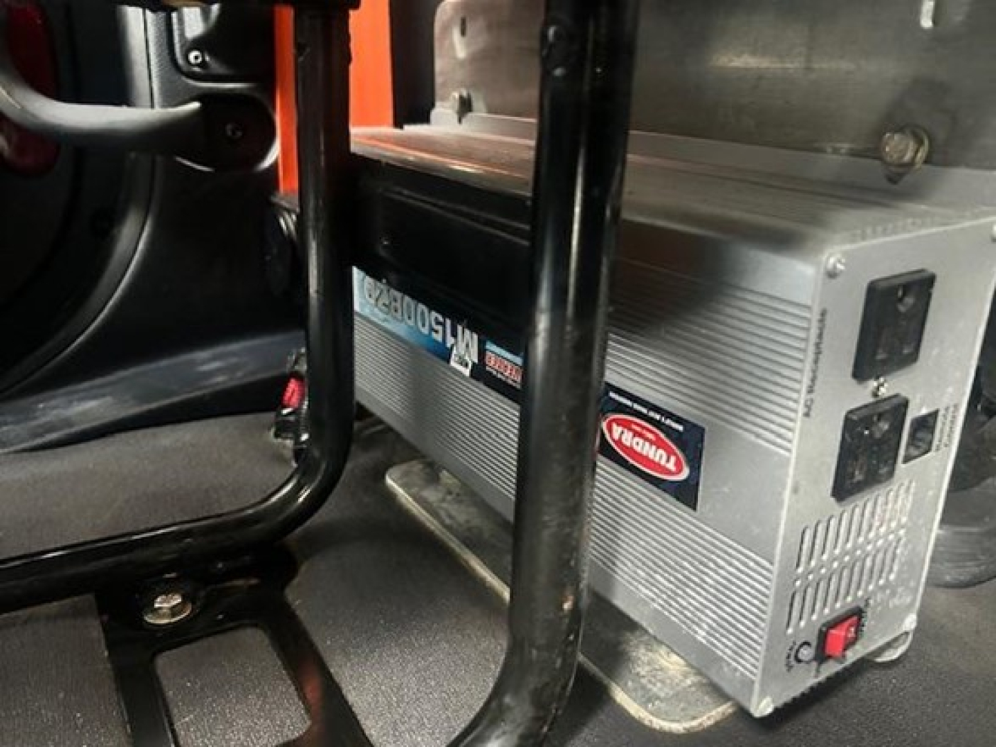 Forklift-like view inside a Freightliner Cascadia cab showing a Tripp Lite power inverter with brand label, placed on floor under seat.