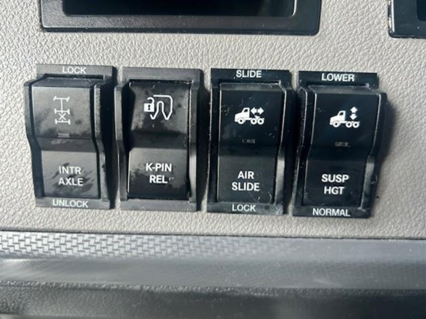 Panel of dashboard switches in a Freightliner Cascadia showing LOCK, SLIDE, LOWER controls with icons for intr axles, K-PIN REL, AIR SLIDE, SUSP HGT, and NORMAL, plus a portion of a digital display above.