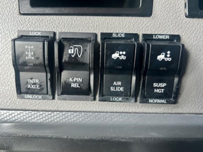 Panel of dashboard switches in a Freightliner Cascadia showing LOCK, SLIDE, LOWER controls with icons for intr axles, K-PIN REL, AIR SLIDE, SUSP HGT, and NORMAL, plus a portion of a digital display above.