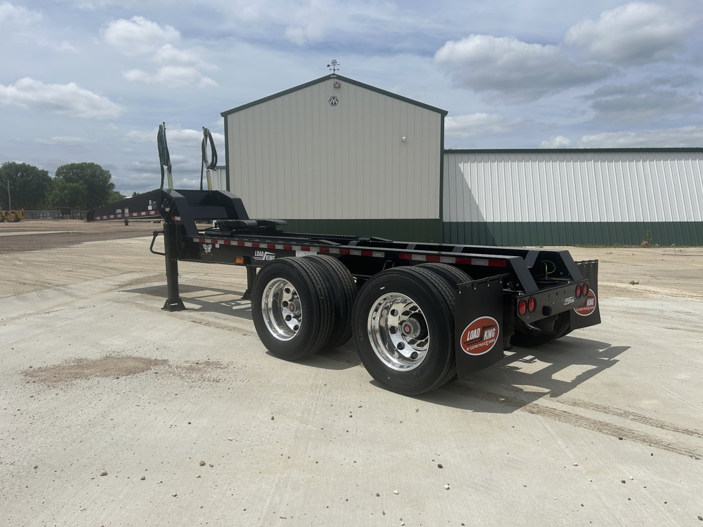 Load King HD702G trailer showing dual rear axles with black fenders and red circular warning decals; metal frame, wheel hubs, and a side marker light visible.