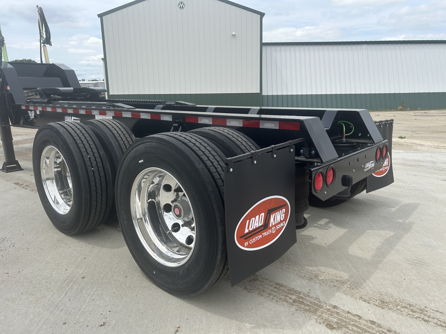 Load King HD702G trailer showing dual rear axles with large chrome rims and black mud flaps; rear frame and deck visible with reflective tape.
