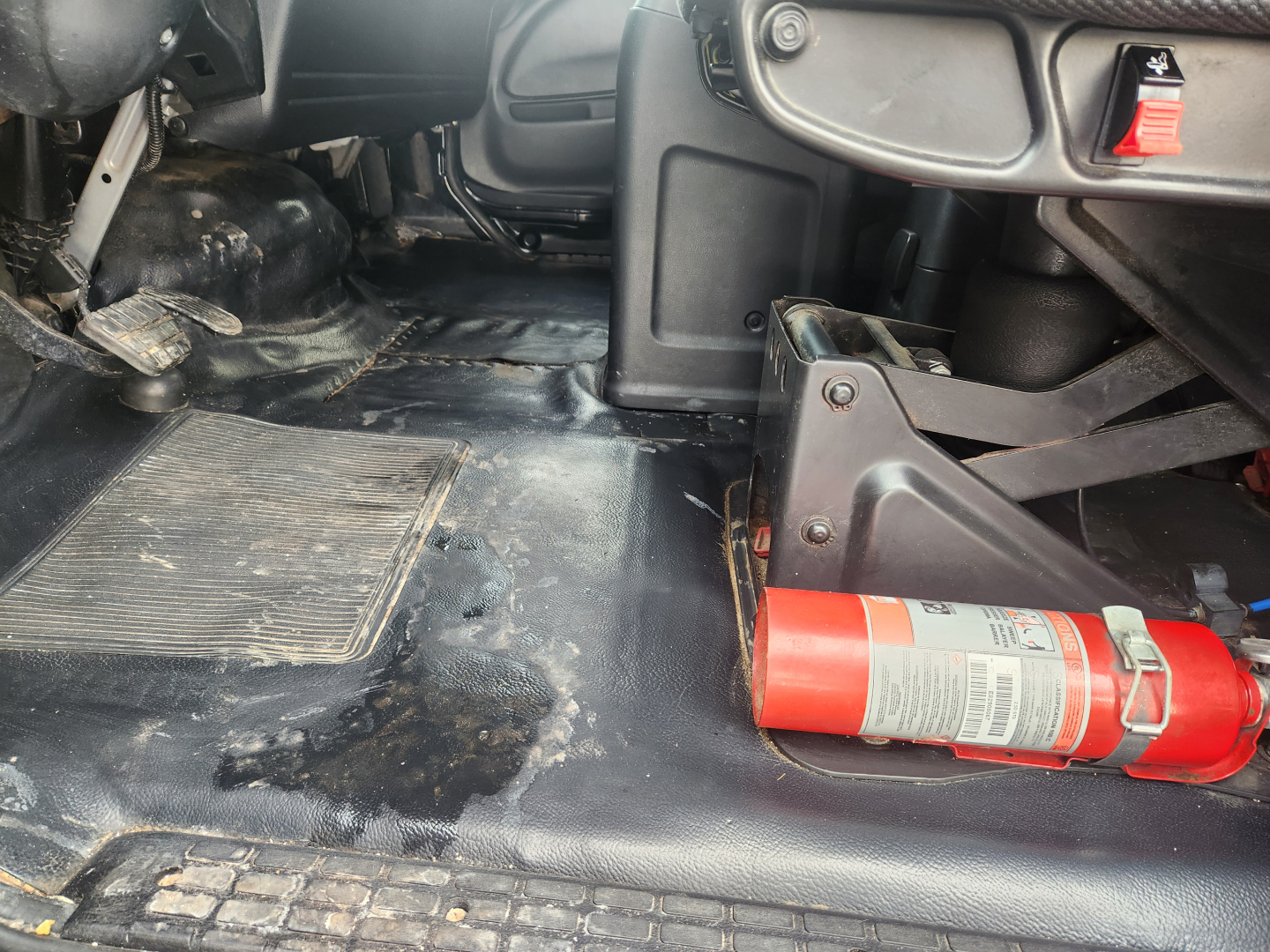 Interior cabin floor area of a 2020 Hino 268A with a red fire extinguisher mounted near the driver’s seat and various panels; dirt and wear on the floor mat and metal floor.