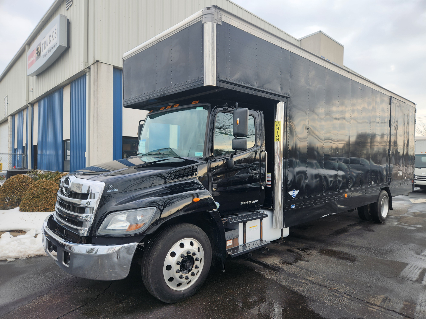 Hino 268A moving van truck in showroom lot with black box body and open side cab showing step access.