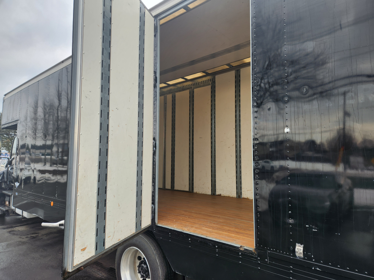 Truck trailer interior with open rear ramp door showing empty plywood floor and sidewall metal studs; black exterior with riveted panels.