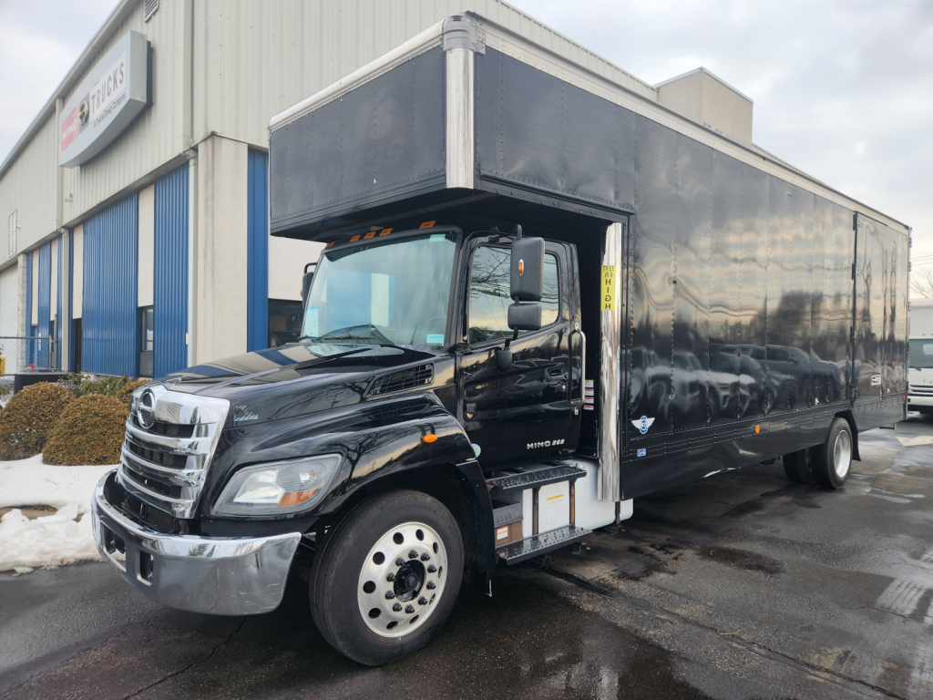 Hino 268A moving van truck in showroom lot with black box body and open side cab showing step access.