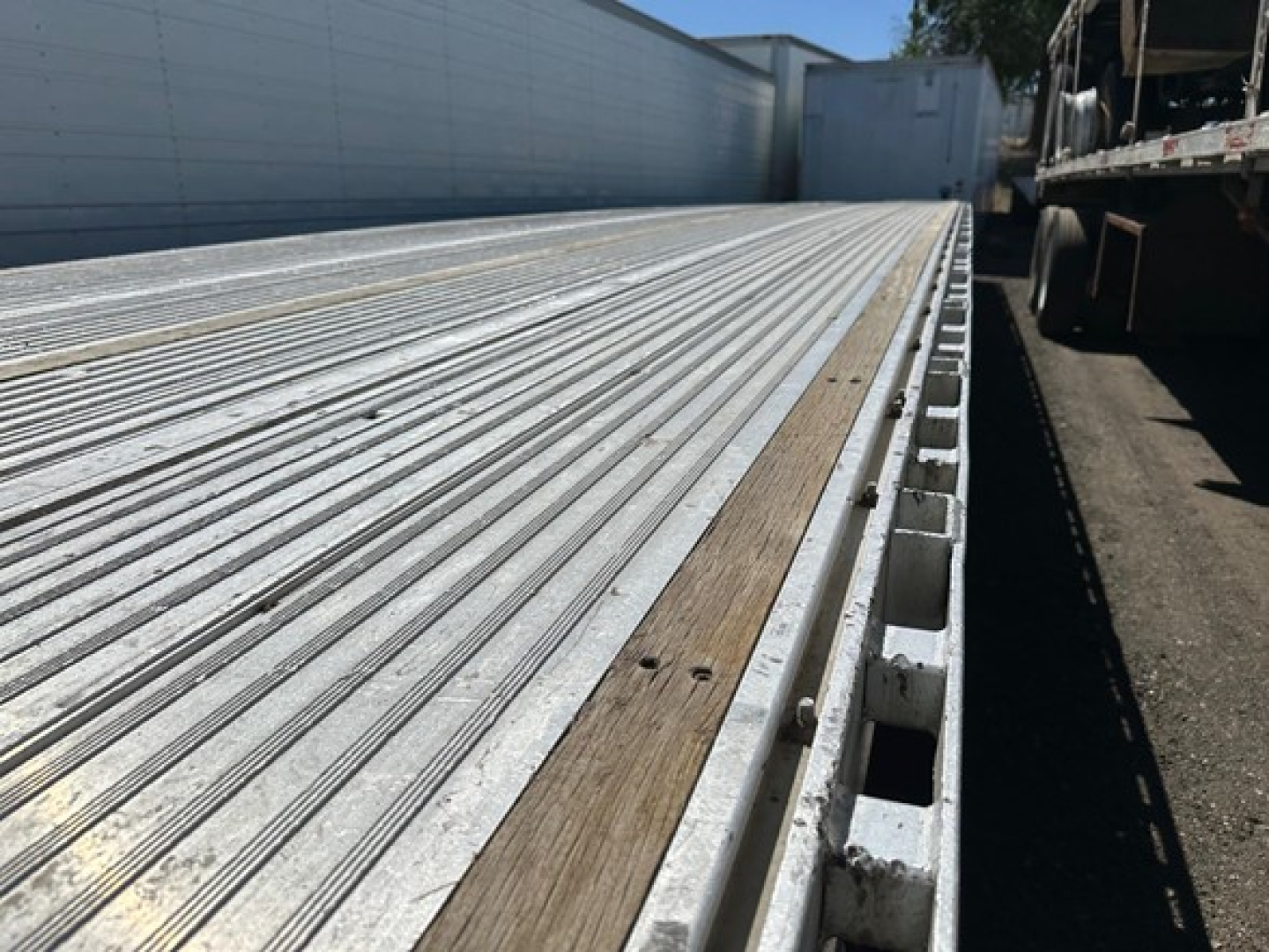 All-aluminum drop deck trailer bed with ribbed aluminum surface and wood plank edge, perspective view along the length.
