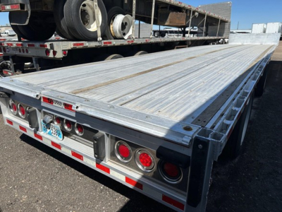 All-aluminum drop deck trailer bed with rear lights and reflective tape; flat silver surface with ribbed deck and side stake pockets; rear bumper and license plate visible.