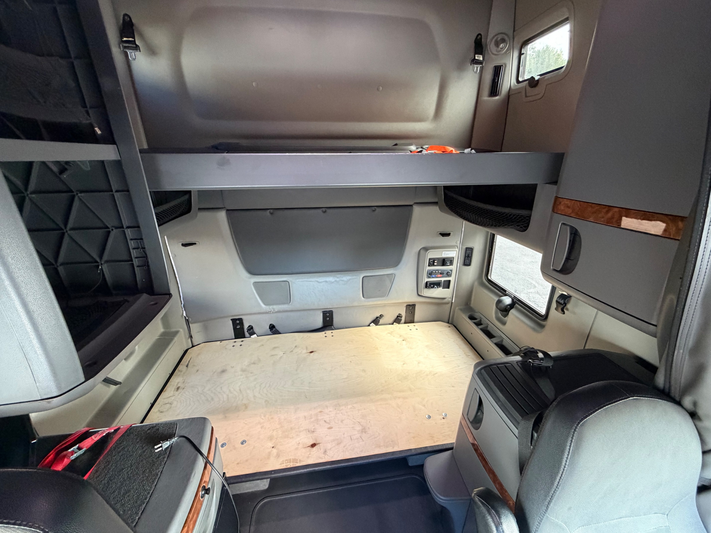 Interior sleeper cab showing bunk bed platform above the cab area and storage beside windshield, with a bare wooden floor and control panels on the side.