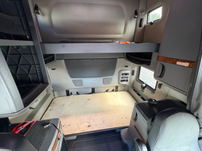 Interior sleeper cab showing bunk bed platform above the cab area and storage beside windshield, with a bare wooden floor and control panels on the side.
