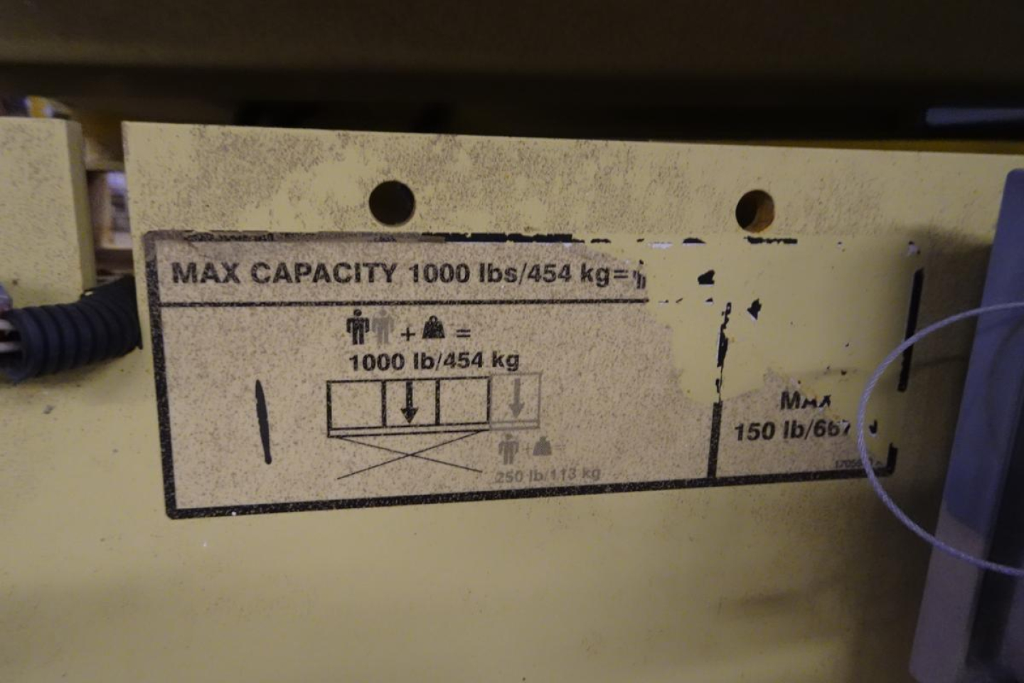 Label shows capacity info: MAX CAPACITY 1000 lbs/454 kg. Diagrams with arrows and weight 1000 lb/454 kg. Plus MAA 150 lb/667? on right side. Some vertical arrows and pictograms. 2646ES scissor lift identification label.