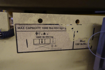 Label shows capacity info: MAX CAPACITY 1000 lbs/454 kg. Diagrams with arrows and weight 1000 lb/454 kg. Plus MAA 150 lb/667? on right side. Some vertical arrows and pictograms. 2646ES scissor lift identification label.
