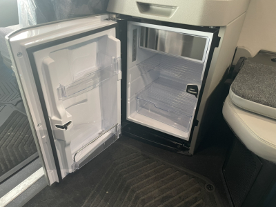 Volvo VNL64T860 interior showing small adjacent refrigerator/freezer open with empty shelves and door storage, compact layout between driver's seat and floor mats.