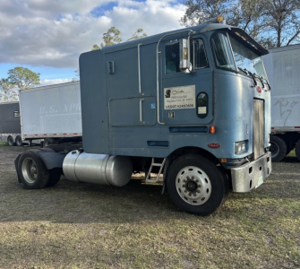 Peterbilt cabover truck in blue with sleeper cab, single rear axle, fuel tanks, and a large grille; US DOT placard and bumper details visible.