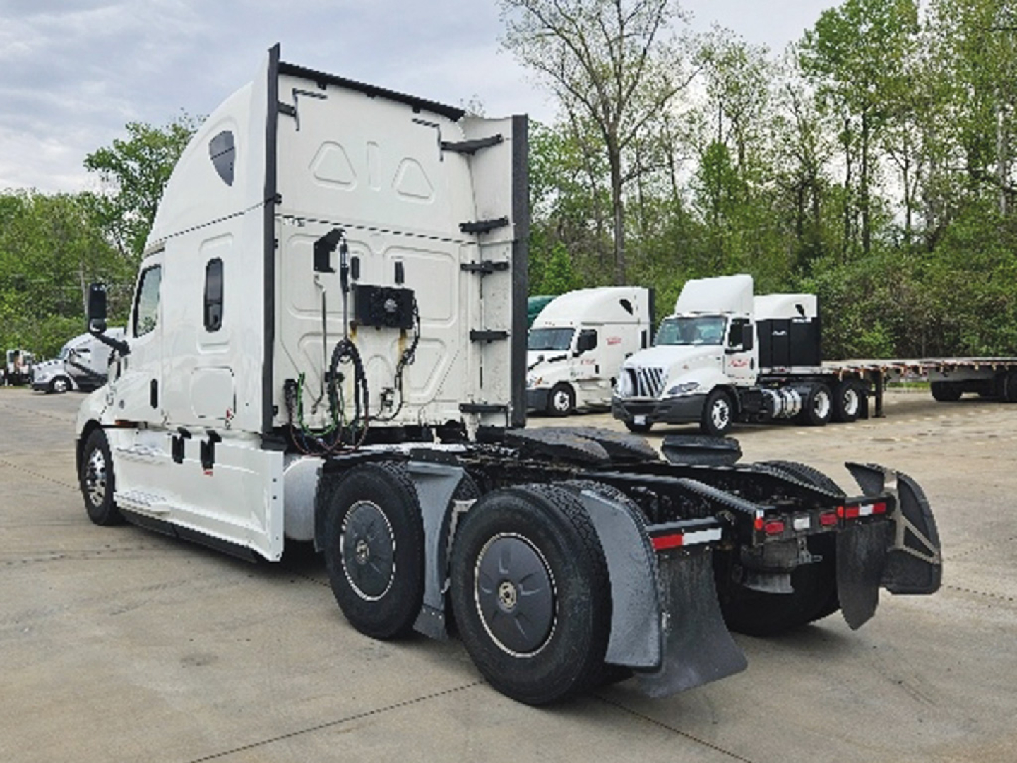 Freightliner Cascadia126 tractor head viewed from rear-left, showing fifth wheel area and rear dual wheels, white cab, parking lot of trucks in background.