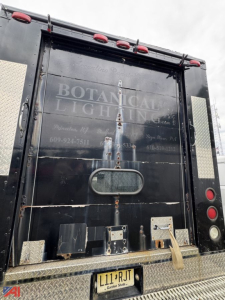 Isuzu NPR box truck rear showing service doors, rusted hardware, diamond plate step bumper, and faded text 'BOTANICAL LIGHTING' on black rear doors.