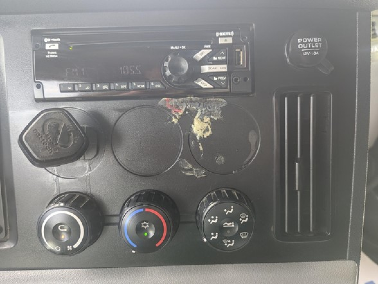 Dashboard panel of a Freightliner Cascadia 126 truck showing climate controls, cigarette lighter, and media head unit with a damaged circular component area.