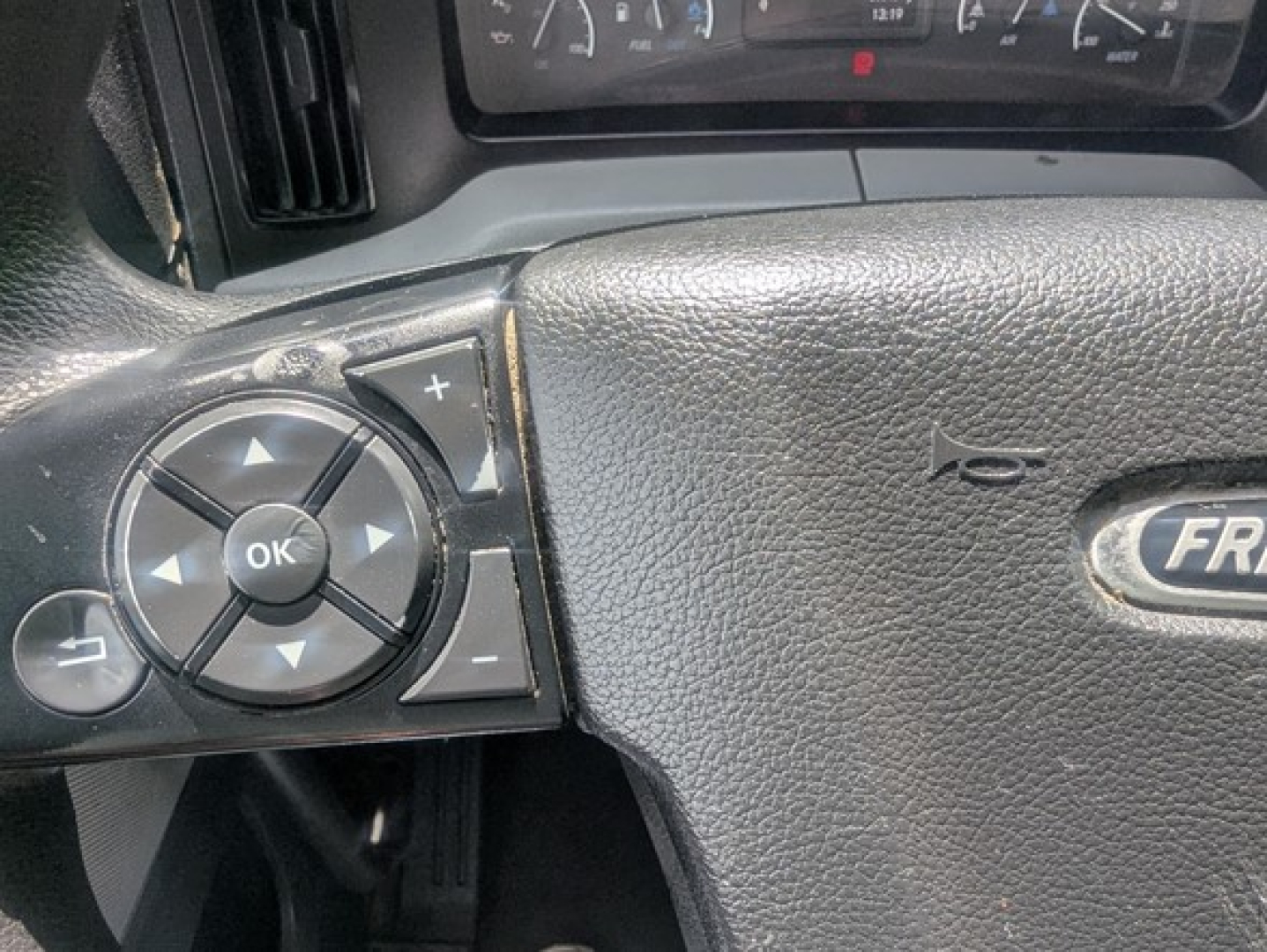 Steering wheel close-up of a Freightliner Cascadia showing control pad with OK button and directional arrows.