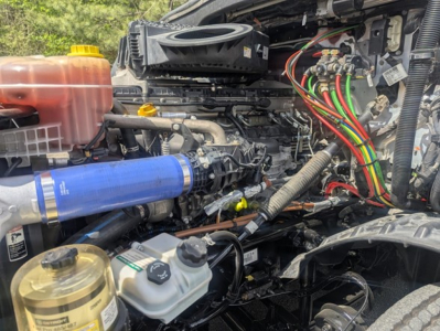 Freightliner Cascadia 126 engine bay with labeled hoses and reservoirs; visible components include coolant reservoir, blue cold air intake, brake fluid reservoir, power steering/auxiliary lines, and assorted wiring harnesses.