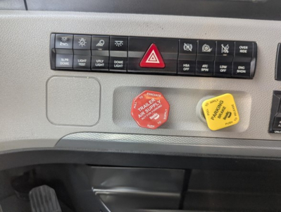 Dashboard of a Freightliner Cascadia showing hazard light switch, air suspension controls, and two colored caps labeled 'TRAILER AIR SUPPLY' red and yellow. 