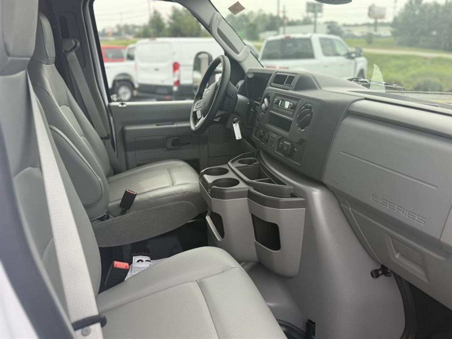 Ford E-450 interior view showing front seats, steering wheel, center console with cup holders, dashboard, and gear area. Light gray upholstery and plastic trim.