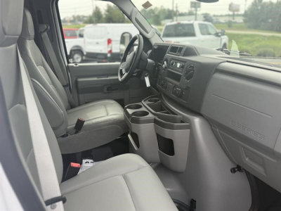 Ford E-450 interior view showing front seats, steering wheel, center console with cup holders, dashboard, and gear area. Light gray upholstery and plastic trim.
