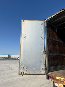 Trailer van rear door open showing interior wooden slat walls and metal frame, with exterior silver door and rust spots at edges.