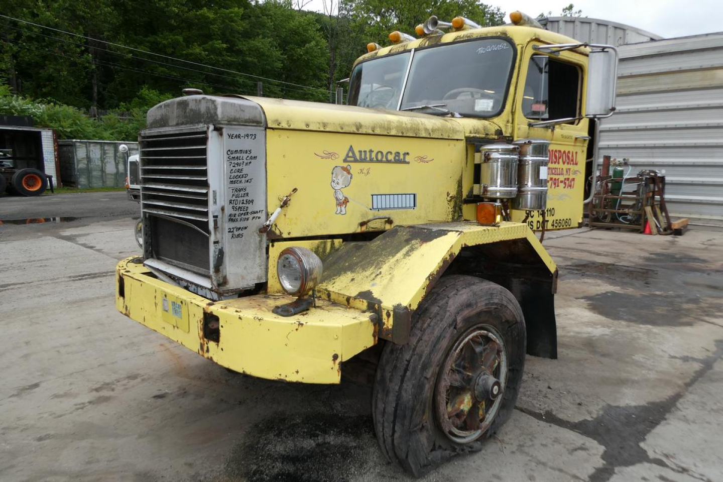 Autocar day cab truck from 1973 with yellow paint, front grille, bumper, and stickered cab showing Autocar branding and service decals; single front wheel visible; weathered body.