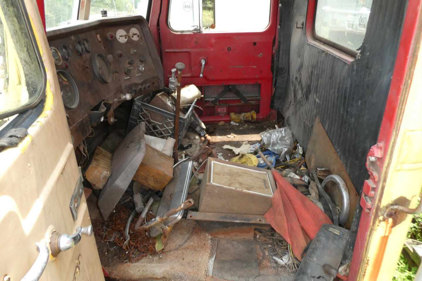 Interior of a worn truck cabin showing a cluttered floor with metal parts, red walls, and a steering wheel on the left side.