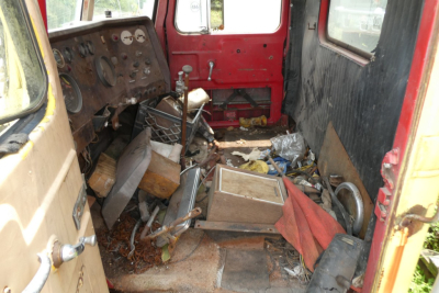 Interior of a worn truck cabin showing a cluttered floor with metal parts, red walls, and a steering wheel on the left side.