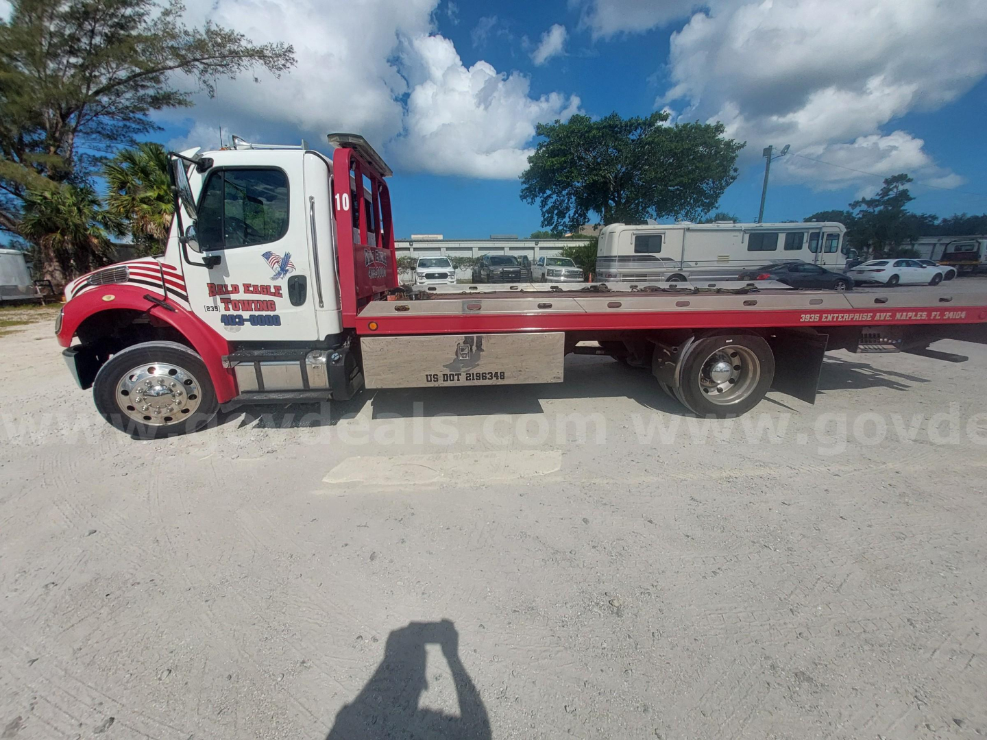 Flatbed tow truck side view showing Freightliner M2 cab, red and white color, lettering on door, long flatbed with wheelset, clear sky background.