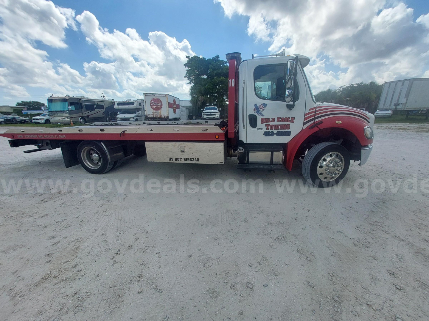 Tow truck side view showing Freightliner M2 with flatbed, company decals on cab door, and wheel wells. Visible text includes company name and contact on door and side steps.